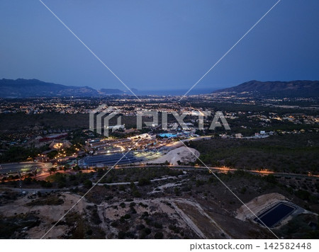 Coastal town twilight panorama with illuminated streets, residential districts, distant mountains, and calm sea horizon beneath deep blue evening sky, with twilight, aerial, panorama, coastal detail Coastal town twilight panorama with illuminated streets, residential districts, distant mountains, and calm sea horizon beneath deep blue evening sky, with twilight, aerial, panorama, coastal detail 142582448