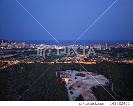 Coastal city skyline at dusk with illuminated towers, shoreline resort district, calm sea, distant island, golf course, and warm blue hour light over waterfront Coastal city skyline at dusk with illuminated towers, shoreline resort district, calm sea, distant island, golf course, and warm blue hour light over waterfront 142582451