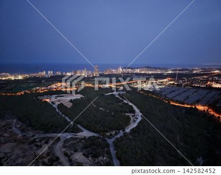 Coastal city skyline at dusk with illuminated highrise towers, shoreline sea views, distant island horizon, winding roads, suburban districts, forested foreground and bluehour atmosphere Coastal city skyline at dusk with illuminated highrise towers, shoreline sea views, distant island horizon, winding roads, suburban districts, forested foreground and bluehour atmosphere 142582452