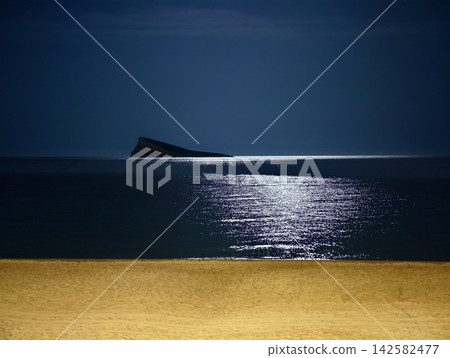 Moonlit seascape with distant rocky islet, calm reflective water, dark blue sky, soft shoreline sand, and quiet horizon creating minimalist nighttime coastal mood Moonlit seascape with distant rocky islet, calm reflective water, dark blue sky, soft shoreline sand, and quiet horizon creating minimalist nighttime coastal mood 142582477