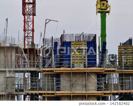 Concrete building under construction with scaffolding, formwork, rebar, and tower cranes rising beside unfinished floors against cloudy sky in busy urban worksite Concrete building under construction with scaffolding, formwork, rebar, and tower cranes rising beside unfinished floors against cloudy sky in busy urban worksite 142582482