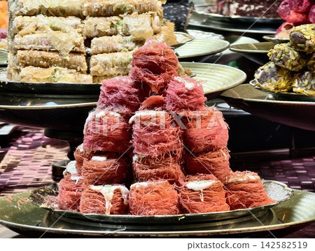 traditional middle eastern desserts stacked and displayed on trays in a culinary market stall with kunefe and baklava, red and beige colors, delicious and inviting mood, close-up from above. 142582519