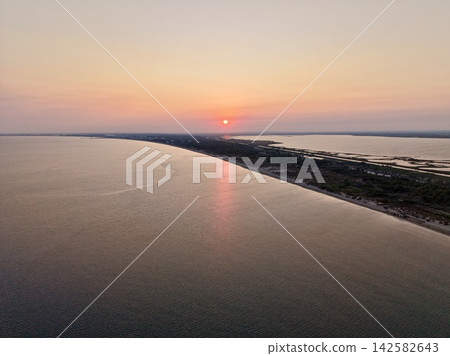 Sunset over a coastal landscape setting sun, in coastal area with sandy beach and vegetation, with sun, ocean, beach, vegetation, from a aerial view perspective. 142582643
