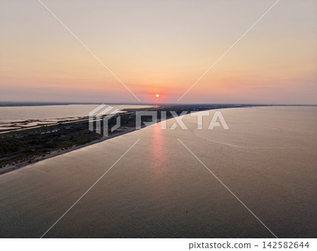 peaceful sunset over sandy beach with green vegetation and calm water from aerial view above the coastal area with orange and pink hues in the sky and sand below. peaceful sunset over sandy beach with green vegetation and calm water from aerial view above the coastal area with orange and pink hues in the sky and sand below. 142582644