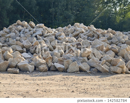 Large outdoor pile of gray rocks and boulders beside dirt ground with forest backdrop and sunlight, with rock, boulder, stone, gravel detail, showing pile, outdoor, yard, construction context Large outdoor pile of gray rocks and boulders beside dirt ground with forest backdrop and sunlight, with rock, boulder, stone, gravel detail, showing pile, outdoor, yard, construction context 142582784