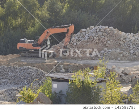 Orange excavator at rocky quarry site with large rubble pile, heavy machinery, lush green trees in background, dusty ground, exposed hydraulic arm, protective cab guard, construction equipment on Orange excavator at rocky quarry site with large rubble pile, heavy machinery, lush green trees in background, dusty ground, exposed hydraulic arm, protective cab guard, construction equipment on 142582785