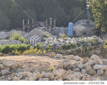 Wide view construction site with rock piles, pillars, scaffolding, blue storage tank beside building amid forest, with rock, quarry, construction, site detail, showing industrial, equipment 142582786