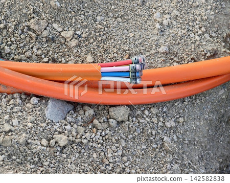 Coiled orange electrical cables lie on rough gravel and rocks at construction site, exposed hoses and connectors among dirt and debris, heavy duty piping and multicolor cables on ground Coiled orange electrical cables lie on rough gravel and rocks at construction site, exposed hoses and connectors among dirt and debris, heavy duty piping and multicolor cables on ground 142582838