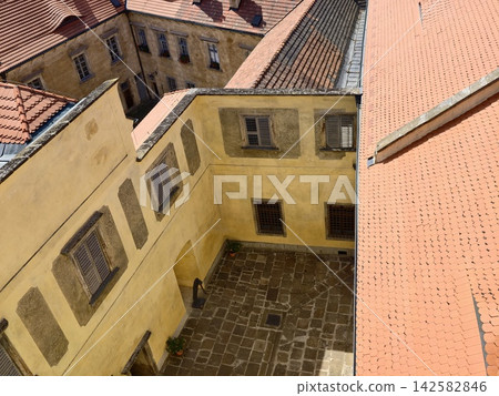 Aerial view of sunlit European courtyard surrounded by warm-toned tiled rooftops and yellow sandstone walls, with courtyard, rooftop, tile, terracotta detail, showing bellow, yellow, building Aerial view of sunlit European courtyard surrounded by warm-toned tiled rooftops and yellow sandstone walls, with courtyard, rooftop, tile, terracotta detail, showing bellow, yellow, building 142582846