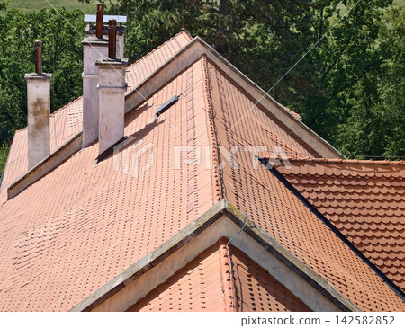 Terracotta tiled roof with chimneys under bright daylight and lush green trees forms architectural rooftop scene, with roof, tile, terracotta, rooftop detail, showing chimney, ridge, slope, pattern Terracotta tiled roof with chimneys under bright daylight and lush green trees forms architectural rooftop scene, with roof, tile, terracotta, rooftop detail, showing chimney, ridge, slope, pattern 142582852