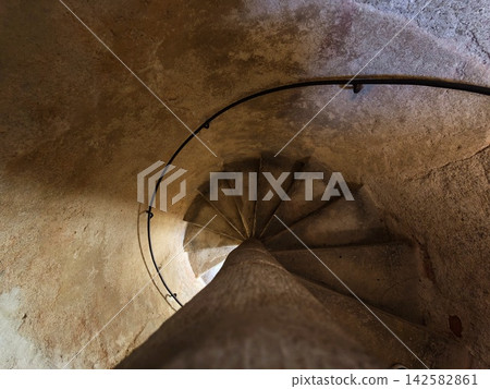 Spiral staircase curves inward inside aged stone tower, weathered walls and iron railing tracing circular path under dim light, with spiral, staircase, stone, tower detail Spiral staircase curves inward inside aged stone tower, weathered walls and iron railing tracing circular path under dim light, with spiral, staircase, stone, tower detail 142582861