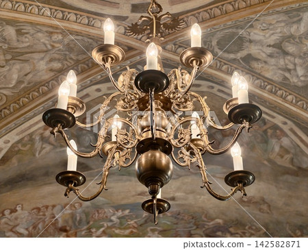 Ornate bronze chandelier with candle-shaped bulbs hanging above richly painted historic ceiling mural showing intricate metalwork and classical lighting in elegant interior Ornate bronze chandelier with candle-shaped bulbs hanging above richly painted historic ceiling mural showing intricate metalwork and classical lighting in elegant interior 142582871