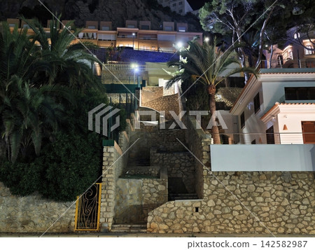 Night view of illuminated hillside residential, complex with stone staircases, palm trees, balconies, and warm exterior lighting along terraced stone walls, with night, stair, staircase, stone detail Night view of illuminated hillside residential, complex with stone staircases, palm trees, balconies, and warm exterior lighting along terraced stone walls, with night, stair, staircase, stone detail 142582987
