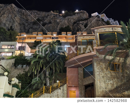 Night view hillside rocks illuminated terrace buildings along winding yellow railing stairway under dark sky overlooking coastal hillside resort, with night, hillside, rock, cliff detail 142582988