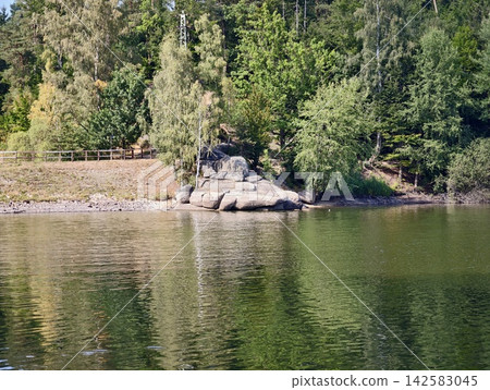 Sunlit lakeside shoreline featuring large rocks and boulders, dense pine forest, calm reflective water, green ripples, distant wooden railing, and tranquil natural scenery by shore in bright daylight Sunlit lakeside shoreline featuring large rocks and boulders, dense pine forest, calm reflective water, green ripples, distant wooden railing, and tranquil natural scenery by shore in bright daylight 142583045