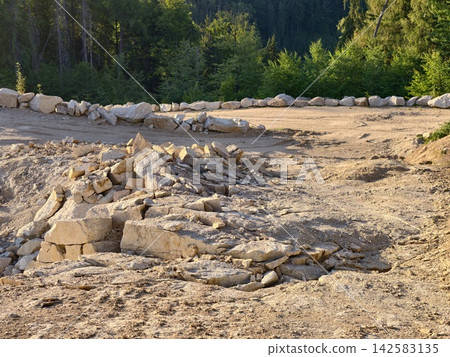 Rugged construction site with stone retaining wall dirt ramp rocky terrain and forest backdrop under daylight capturing outdoor earthwork and natural boundary across open landscape Rugged construction site with stone retaining wall dirt ramp rocky terrain and forest backdrop under daylight capturing outdoor earthwork and natural boundary across open landscape 142583135