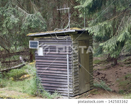 Weathered wooden shed beside tall pine trees in quiet forest, moss-covered roof, mounted antenna, rough rural outbuilding details and muted natural light creating secluded countryside mood Weathered wooden shed beside tall pine trees in quiet forest, moss-covered roof, mounted antenna, rough rural outbuilding details and muted natural light creating secluded countryside mood 142583162