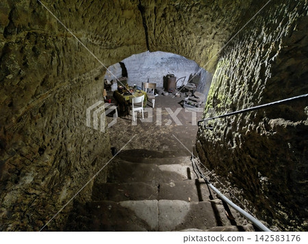 Interior view mossy stone cave dining area with worn wooden chairs metal stove round table set with dishes cups kettle under curved rock arch rustic abandoned camp scene Interior view mossy stone cave dining area with worn wooden chairs metal stove round table set with dishes cups kettle under curved rock arch rustic abandoned camp scene 142583176