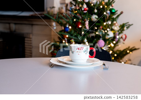 Festive Cup and Saucer on White Table by Christmas Tree. A festive patterned cup and saucer on a white table in front of a glowing Christmas tree with a fireplace behind. Warm bokeh lights create Festive Cup and Saucer on White Table by Christmas Tree. A festive patterned cup and saucer on a white table in front of a glowing Christmas tree with a fireplace behind. Warm bokeh lights create 142583258
