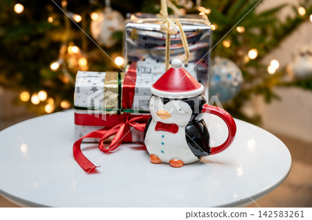 Cute Penguin Santa Mug and Wrapped Gifts by Christmas Tree. A cute penguin mug in a Santa hat sits beside festive wrapped gifts with red ribbons on a white table in front of a glowing Christmas tree.  142583261