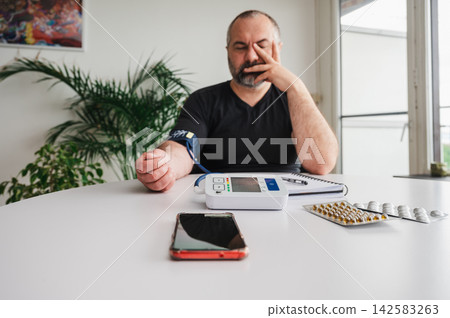 Man Recording Blood Pressure Reading in Notebook Vertical. Vertical photo of a mature man writing down his blood pressure reading in a notebook beside a home monitor at a table, illustrating health Man Recording Blood Pressure Reading in Notebook Vertical. Vertical photo of a mature man writing down his blood pressure reading in a notebook beside a home monitor at a table, illustrating health 142583263