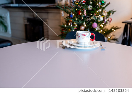 Festive Mug and French Press Tea with Candle by Tree. A festive mug, a glass French press of tea and a lit tea light on a white table before a glowing Christmas tree. Warm bokeh lights create a cozy Festive Mug and French Press Tea with Candle by Tree. A festive mug, a glass French press of tea and a lit tea light on a white table before a glowing Christmas tree. Warm bokeh lights create a cozy 142583264