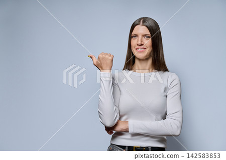 Portrait of young woman pointing thumb to copy space on grey background Portrait of young woman pointing thumb to copy space on grey background 142583853