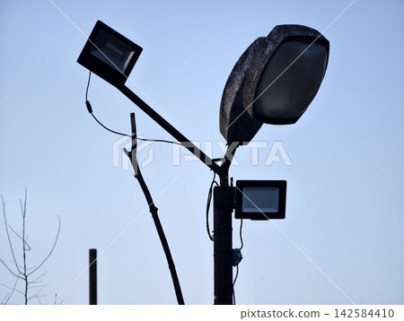 City lamp with floodlights. Modern urban lighting fixture with floodlights and city backdrop City lamp with floodlights. Modern urban lighting fixture with floodlights and city backdrop 142584410