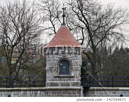 Old stone gatehouse along dam pathway under cloudy sky with iron railing and tranquil water edge Old stone gatehouse along dam pathway under cloudy sky with iron railing and tranquil water edge 142584671