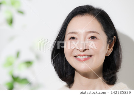 A woman in her 50s smiles naturally, looking directly at the camera against a backdrop of fresh spring greenery, showcasing her translucent, radiant beauty and healthy, beautiful skin. A woman in her 50s smiles naturally, looking directly at the camera against a backdrop of fresh spring greenery, showcasing her translucent, radiant beauty and healthy, beautiful skin. 142585829