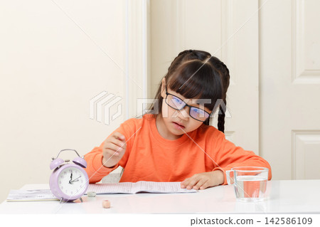 An Asian girl doing her homework looks at the alarm clock, expressing the concept of being late or time pressure. An Asian girl doing her homework looks at the alarm clock, expressing the concept of being late or time pressure. 142586109