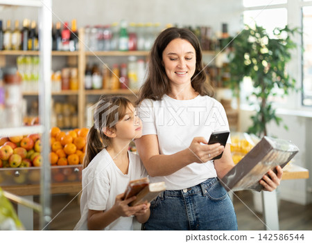 Girl with mother customers at shop scans QR code on sweets package using phone camera Girl with mother customers at shop scans QR code on sweets package using phone camera 142586544
