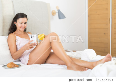 Woman drinking morning coffee and looking at smartphone screen in bedroom Woman drinking morning coffee and looking at smartphone screen in bedroom 142586793