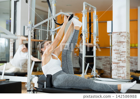 Girl works out in gym and performs set of exercises to stretch leg muscles with help of reformer Girl works out in gym and performs set of exercises to stretch leg muscles with help of reformer 142586988