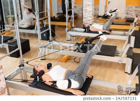 Girl works out in gym and performs set of exercises to stretch leg muscles with help of reformer Girl works out in gym and performs set of exercises to stretch leg muscles with help of reformer 142587131