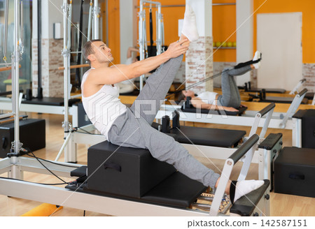 Man works out in gym and performs set of exercises to stretch leg muscles with help of reformer Man works out in gym and performs set of exercises to stretch leg muscles with help of reformer 142587151