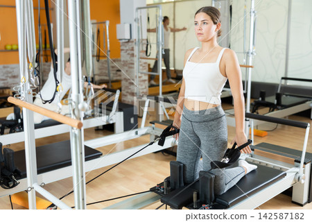 Girl in gym with help of rope reformer performs exercises to strengthen muscles of her arms. Girl in gym with help of rope reformer performs exercises to strengthen muscles of her arms. 142587182
