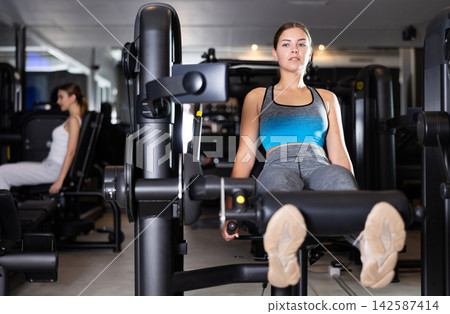 Sportive young girl training on leg extension machine in gym Sportive young girl training on leg extension machine in gym 142587414