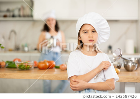 Little girl in chefs hat and white T-shirt stands in kitchen near table Little girl in chefs hat and white T-shirt stands in kitchen near table 142587426