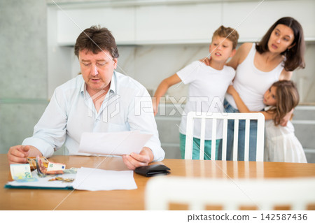 Worried adult man examining documents and counting money at table with family Worried adult man examining documents and counting money at table with family 142587436