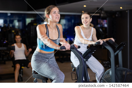 During classes on air bike, girls chat sweetly and smile During classes on air bike, girls chat sweetly and smile 142587464