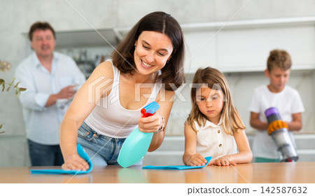 Mom and little daughter are dusting the table. Father and son cleaning the room in background Mom and little daughter are dusting the table. Father and son cleaning the room in background 142587632
