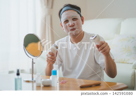 A 40-something man using a facial mask while holding two facial devices in his hands – a men's beauty treatment. 142591080