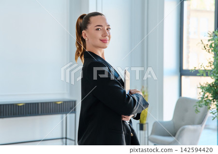 Confident business woman holding closed laptop in modern office Confident business woman holding closed laptop in modern office 142594420
