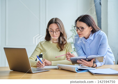 Female teacher explaining task to young student writing at desk Female teacher explaining task to young student writing at desk 142594750