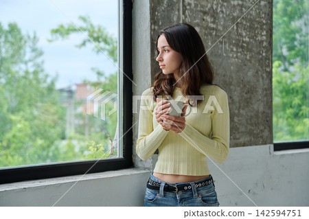 Pensive young woman holding mug standing near window and looking outside Pensive young woman holding mug standing near window and looking outside 142594751