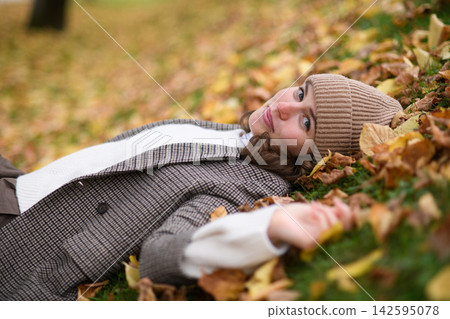 Portrait of a beautiful woman lying in fallen autumn leaves on the lawn. She smiles and looking at camera. Portrait of a beautiful woman lying in fallen autumn leaves on the lawn. She smiles and looking at camera. 142595078