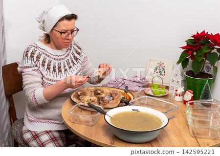 A woman separates boiled meat from bones to prepare kholodets, a jellied meat dish. She works at a wooden table with utensils and ingredients in a cozy kitchen during winter A woman separates boiled meat from bones to prepare kholodets, a jellied meat dish. She works at a wooden table with utensils and ingredients in a cozy kitchen during winter 142595221