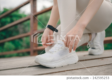 Active athlete runner tying shoelace on sneaker for sport exercise, preparing for outdoor cardiovascular fitness training and running practice on wooden bridge walkway in park 142595253
