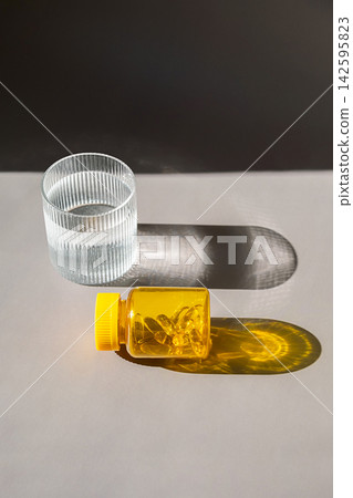 Yellow bottle with gelatin capsules and glass of water on gray table in sunlight with hard shadows. Yellow bottle with gelatin capsules and glass of water on gray table in sunlight with hard shadows. 142595823